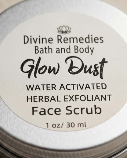 Close-up of a round silver metal tin lid with a cream-colored label reading 'Divine Remedies Bath and Body Glow Dust Water Activated Herbal Exfoliant Face Scrub 1 oz / 30 ml'.