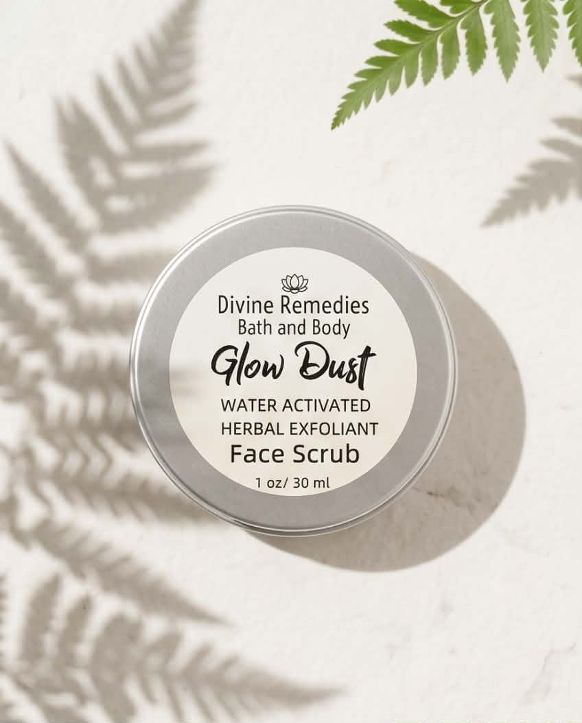 A round silver metal tin of Divine Remedies Glow Dust Face Scrub, photographed from above on a light textured surface with soft fern leaf shadows and a green fern frond in the corner.