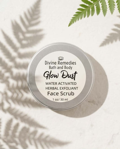 A round silver metal tin of Divine Remedies Glow Dust Face Scrub, photographed from above on a light textured surface with soft fern leaf shadows and a green fern frond in the corner.