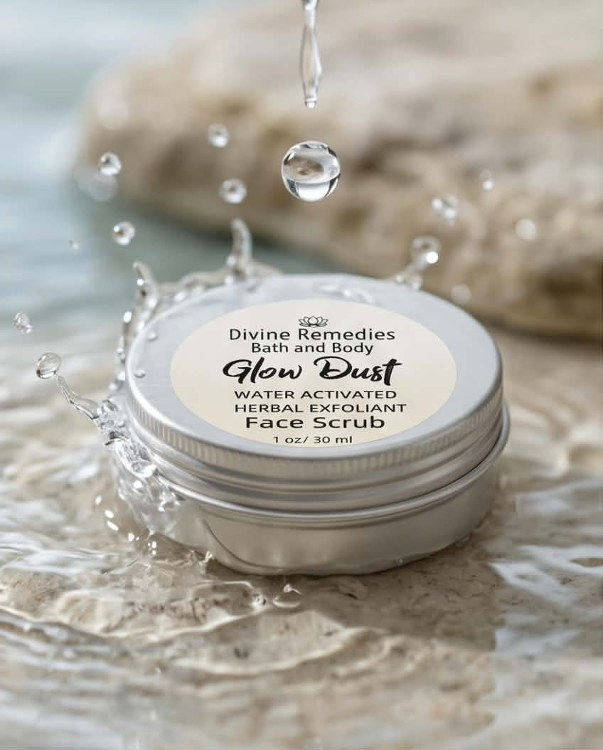 A round metal tin of Divine Remedies Bath and Body Glow Dust Face Scrub sits in shallow water with splashing droplets and a blurred stone background.