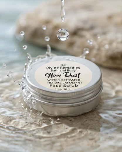 A round metal tin of Divine Remedies Bath and Body Glow Dust Face Scrub sits in shallow water with splashing droplets and a blurred stone background.
